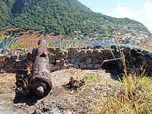 Fort Cachacrou ruins including the remains of a stone wall as well as an old cannon. Below the wall is a body of water, Soufrière Bay.