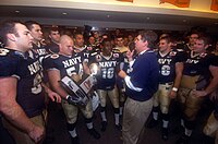 A middle aged man in a blue polo shirt and khaki pants speaks to young men in football uniforms in the locker room. One of the men is holding a trophy.