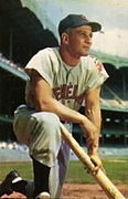 A man in a baseball uniform kneeling on the ground with a baseball bat in hand, with the stadium grandstands in the background.