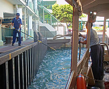 A man at the front of a large boat throws a rope to another man on a nearby pier wearing a blue sailor uniform who is holding a long pole with a hook at the rear to catch it.