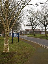 Building with multiple glass windows seen at the end of a tree lined road. Sign says welcome to Norton Radstock College