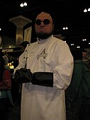 Personne chauve avec une barbe portant la tenue de Hugo Strange, avec la blouse blanche et noire et les lunettes rondes.