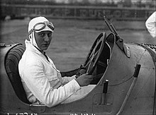 Woman in white suit with goggles on head sitting in racing car