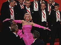 Monroe in Gentlemen Prefer Blondes. She is wearing a shocking pink dress with matching gloves and diamond jewelry, and is surrounded by men in tuxedos