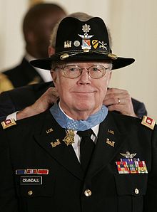 Une photographie en couleurs de Bruce Campbell dans son uniforme militaire avec un chapeau de la cavalerie. Il sourit, et l'on peut voir le président Bush lui accrochant la médaille autour du cou.