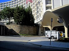 Entrée d'un hôtel blanc dont la façade est courbée. Des arbres et un drapeau américain la décorent au-dessus d'un mur en briques grises.