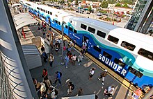 Bahnhof bei Sonnenschein von schräg oben mit Fokus auf Doppelstockzug mit Anschrift SOUNDER und ca. 40 sichtbaren Fahrgästen, die den Fahrgastwechsel vollziehen.