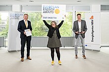 Drei Personen stehen mit etwas Abstand nebeneinander, die Mittlere Person hält ein Schild in die Höhe: European Energy Award – Stadt Emsdetten