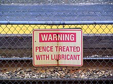 A sign reading "Warning: fence treated with lubricant" on a fence between two railroad tracks