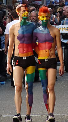 2 jeunes participants à une Gay Pride en Allemagne, tous deux sont vêtus de short et torse nus, leurs cheveux sont teints et ils sont tous deux en partie peints des couleurs de l'arc-en-ciel de leur front à leurs mollets.