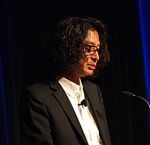 Portrait de Yoshio Sakamoto. Habillé d'une veste de costume noire et d'une chemise blanche, il est pris légèrement sur son profil droit.