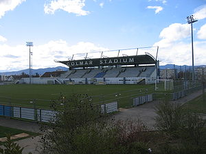Stade de football, sur lequel est écrit Colmar Stadium, vu sous un ciel bleu avec quelques nuages.