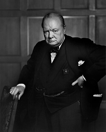 Black-and-white image of Winston Churchill standing with his left hand on his hip and his right hand resting on the back of a chair