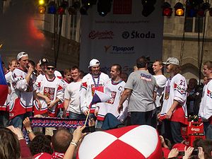 Ice hockey players wearing red and white uniforms stand elevated on a stage facing out towards a crowd of people.