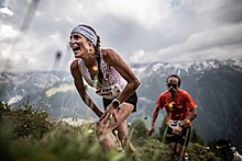 Une femme en habits de sport portant un dossard, les mains sur les genoux, grimpe sur un chemin. Un homme en dossard la suit.