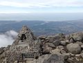 Photo couleur du bord d'un sommet de montagne. Des rochers gris parsèment le sol de terre marron. En bas, à gauche : un monument pyramidal en ciment incrusté de pierre et à l'intérieur évidé. Au deuxième plan : une plaine ; un ciel nuageux, en arrière-plan.