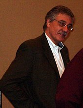 photo en couleur d'un homme aux cheveux gris portant moustache et lunettes