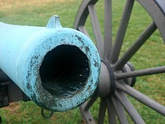 Photo shows the mouth of a cannon that has a pale green patina.