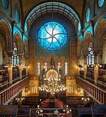 Une grande salle vue de haut. Une grande coupole avec au centre un vitrail bleu. Sur les cotés, des balcon. Vers le bas, un chandelier derrière lequel apparait le sol.