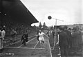 Photographie en noir et blanc d'un coureur à pied à l’arrivée d'une course, encouragé par des hommes qui crient.