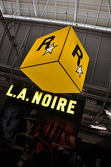 D'un edifici alt penja una gran caixa taronja amb la lletra "R" i una estrella. A sota hi ha un estand de convencions amb el text il·luminat "L.A. NOIRE". La portada del joc es veu vagament sota el logotip.