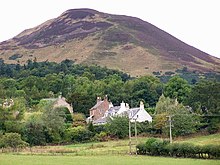 A small cluster of houses sit among trees at the base of a tall bare hill in the background.