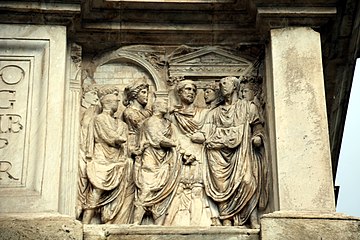 Sur l'attique, à droite de l'inscription : sur le Champ de Mars, devant le temple du dieu, Trajan, avec Hadrien à ses côtés et suivi des licteurs, reçoit les deux consuls, en présence de la déesse Rome.