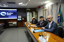 Homem vestido de terno, sentado faz apresentação de slides, enquanto fala.