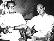 Photographie en noir et blanc montrant deux hommes assis côte à côte sur des chaises en bois. Tous deux portent des uniformes blancs de la marine. L'homme de gauche a les cheveux foncés et légèrement bouclés, ses bras sont croisés et il tient une tasse dans la main droite et porte une montre au poignet gauche. Il ne regarde pas l'objectif. L'autre homme a les cheveux courts et est légèrement chauve. Il a les bras qui reposent sur les accoudoirs de la chaise.