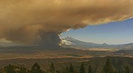 Thick orange-brown smoke blocks half a blue sky, with conifers in the foreground