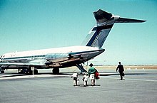 Photographie d'un avion de ligne garé sur le tarmac, avec quatre passagers s'apprêtant à embarquer.