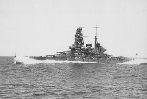 A portside view of Haruna – a large warship with a tall superstructure and two funnels – steaming at full speed in high seas with waves coming over the bow