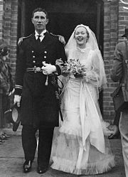 Un jeune couple en habit de mariage se tenant devant un bâtiment. L'homme, qui se tient à gauche, est vêtu d'une tenue de cérémonie navale et tient un chapeau dans sa main droite. La mariée est vêtue d'une robe blanche à voile. Elle sourit et porte un bouquet de fleurs