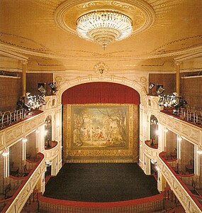 Photographie aux couleurs dominantes rouge et or ; galeries à gauche et à droite, scène au centre, lustre au plafond avec verreries.