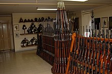Farbfotografie eines Raumes mit Waffen, die in der Mitte senkrecht an Holzgestelle aufgestellt sind. An der hinteren Wand sind altertümliche Blechrüstungen und Helme auf einem Regal aufgereiht.