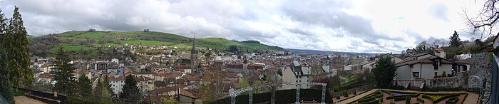 Photo panoramique d'Aurillac, prise du haut d'un des versants de la vallée, d'où l'on voit toutes les maisons de la ville avec l'abbatiale au centre et, à l'arrière-plan, l'autre versant verdoyant qui surplombe la localité. Au-dessus, presque saturé de nuages blancs et parfois gris, un immense ciel malgré tout lumineux.