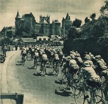 Photo en noir et blanc d'un peloton cycliste se dirigeant vers l'entrée d'une ville avec un château en arrière plan.