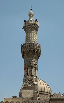 An ornate carved stone minaret, with a carved stone railing around balconies at its center and near its top. The tip of the minaret is a large bulb-shaped stone decoration with a small bulb-shaped metal finial. Behind the minaret part of the top of a dome is visible.