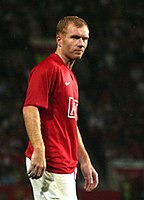 A man with red hair, wearing a red football shirt and white shorts.