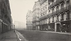 Photo en noir et blanc d'une large rue bordée d'immeubles haussmanniens.