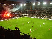 Photo montrant une vue d'ensemble d'un stade, avec des joueurs courant sur la pelouse, et un fumigène allumé dans une tribune.