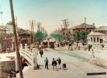 Street scene: People walking over a bridge, with low-rise buildings, street cars, and utility poles leaning crookedly in the background