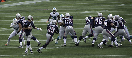 Un joueur court derrière plusieurs joueurs en maillots bleus bloquant leurs adversaires en maillots blancs sur un terrain de football américain.