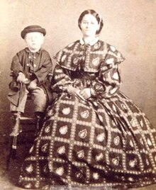 Small blond boy sitting on a stool; to his right a young woman in a full-skirted dress made of fabric with a bold and probably colorful diamond pattern