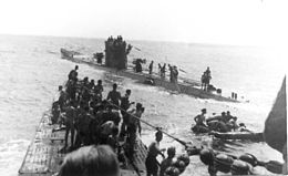 Three-quarter front view over the bow from a submarine conning tower of another submarine with numerous people standing on both submarines, at sea.