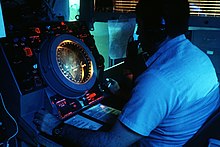 A monitor with a circular appearance is visible. A large man is seated in front of the monitor, and is watching the display with care.