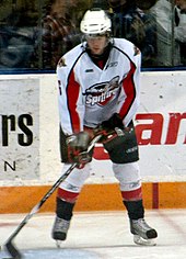 A teenage ice hockey player stickhandling a puck while standing still on the ice. His skates are shoulder-width apart and his eyes are downcast. He wears a white jersey with red and black trim, as well as a white, visored helmet.