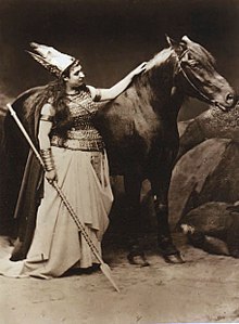Une femme costumée aux côtés d'un cheval sombre, posant sa main sur celui-ci.
