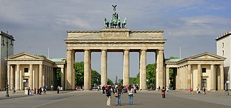 Frontale Farbfotografie vom Brandenburger Tor mit sechs Stützsäulen und einer Quadriga auf dem Dach. An beiden Seiten des Tors stehen zwei kleine klassizistische Gebäude mit Giebelfeldern. Auf dem Vorplatz laufen einige Menschen.