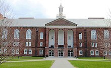 Photographie en couleurs montrant la façade d'un bâtiment en briques rouges, avec un clocher blanc, et trois grandes arcades au-dessus de l'entrée. Devant le bâtiment se trouve un large espace de pelouse.